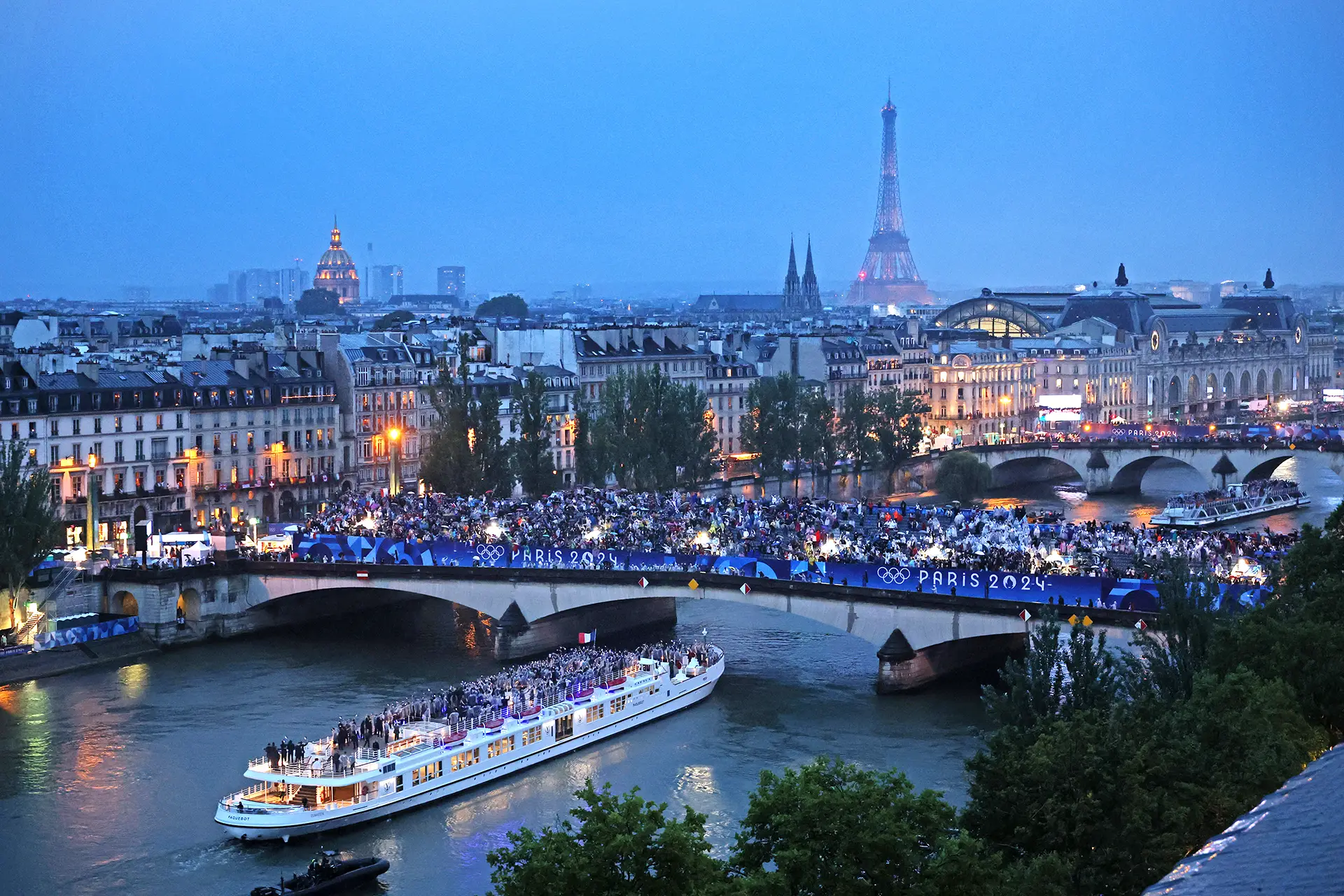 Boats filled with olympic athletes goes under a bridge full of spectators cheering for them, the Eiffel Tower in the background