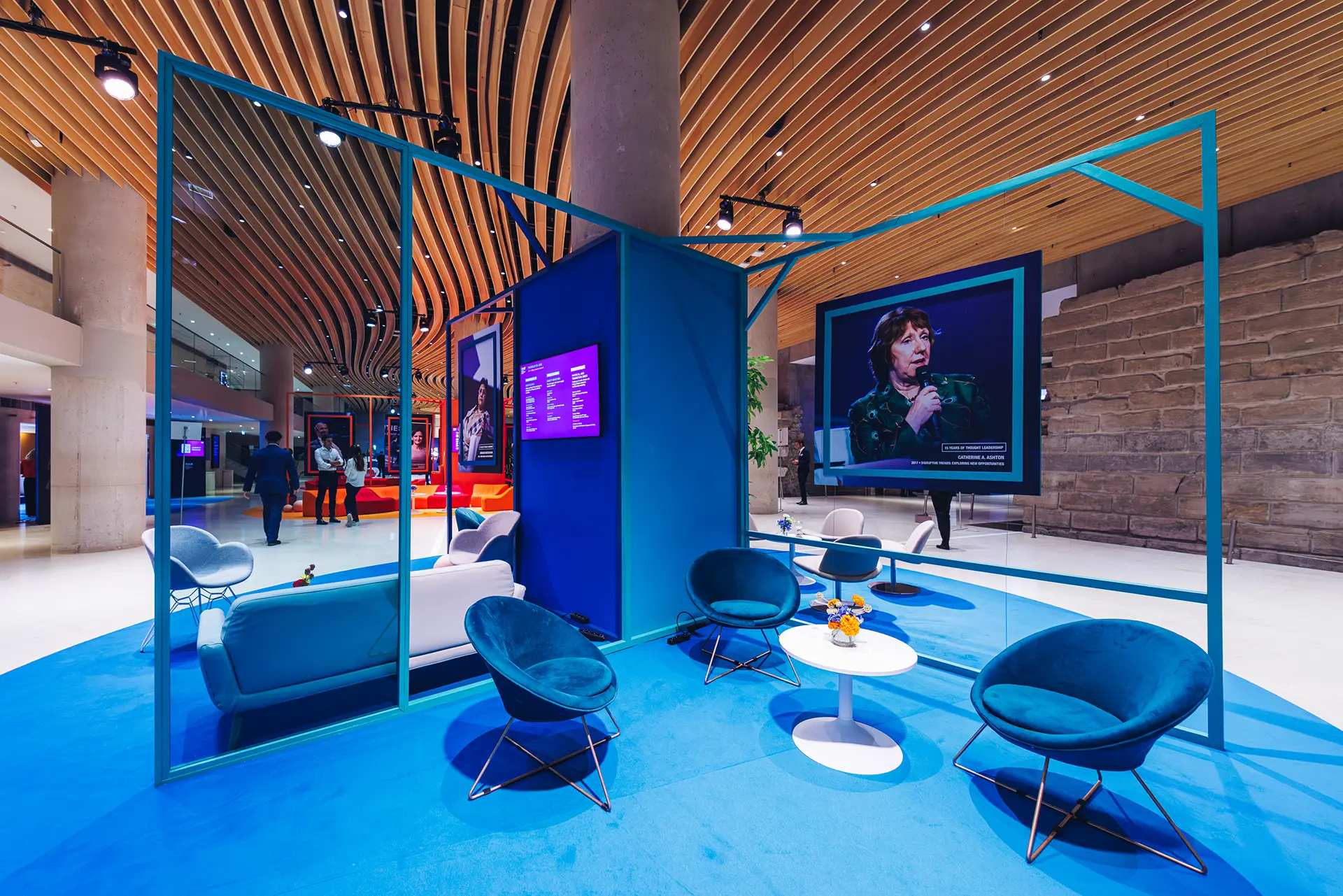 Networking lounge, blue furniture, and panels featuring previous key speakers, such as Catherine A. Ashton in 2017