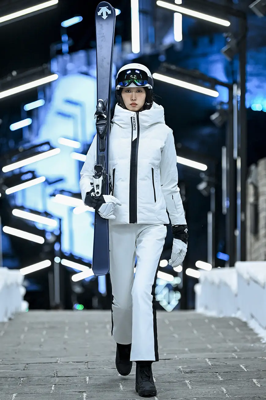 Model on the runway holding a ski pair and wearing white DESCENTE winter sportswear